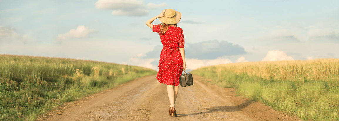 Woman in red dress with suitcase walking down a country road into the horizon.