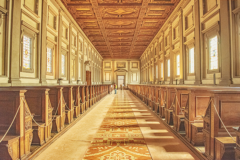 Photo of the interior of Michelangelo's Laurentian Library in Florence. The centre aisle is tiled and wooden reading desks are in rows down either side. It has a golden glow from the timber and the light. This is an authentic experience for travel writing.