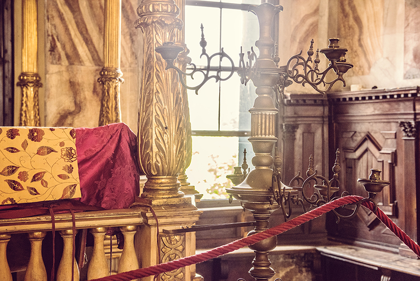 Photo of the interior of the Spanish Synagogue in the Jewish ghetto in Venice. The carved wood is painted gold and a large candelabra sits with no candles leaning towards the window. This quiet place is an example of an authentic travel experience.