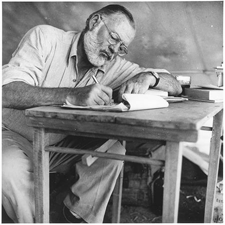 Photograph of Ernest Hemingway sitting at a table writing while at his campsite in Kenya. How to make your writing more powerful.